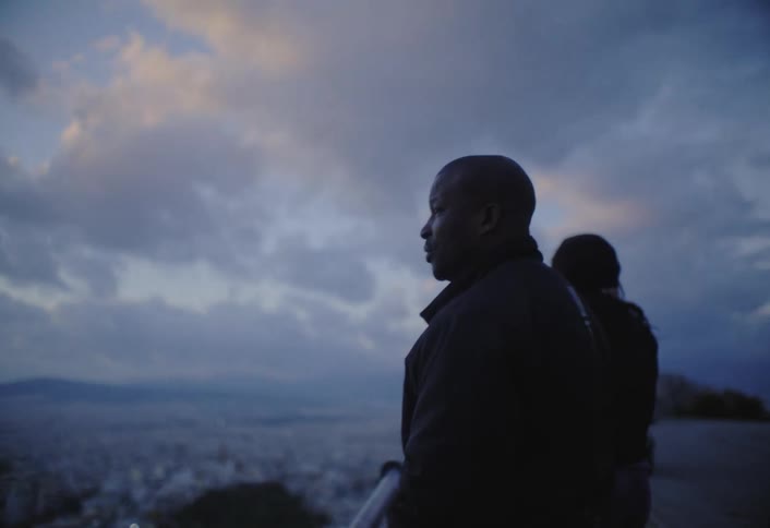 The video shows the silhouettes of two people from behind facing a cloudy sky the horizon reveals a distant city or landscape under the dramatic cloud cover