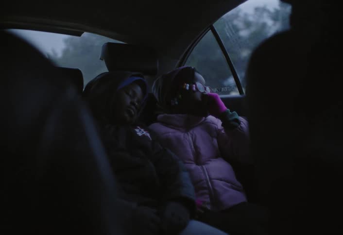 Two kids are sitting side by side in the back seat of a car wearing winter jackets as they go on a road trip at midday