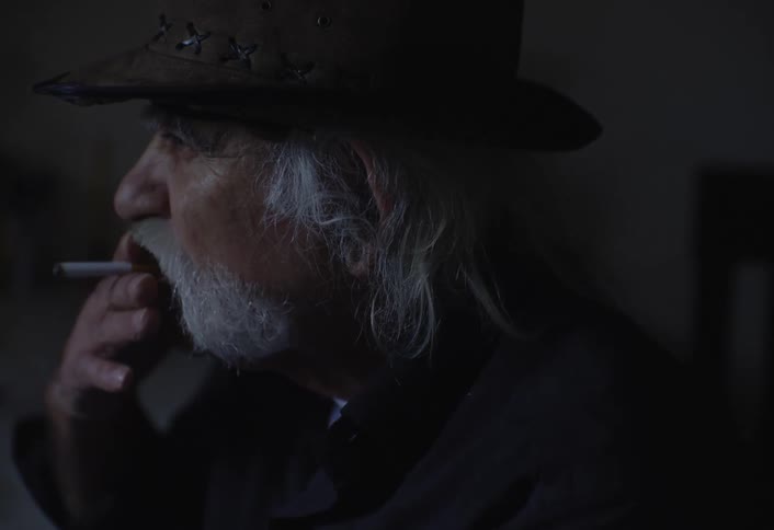 An elderly man in a hat smokes a cigar indoors while speaking