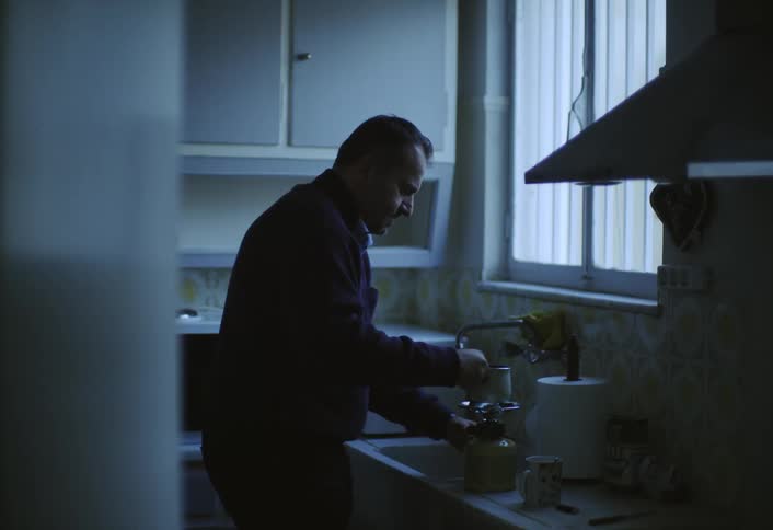 A person stands in a dimly lit kitchen seemingly focused on making coffee using a moka pot on a stovetop the scene is bathed in a cool subdued light possibly from a window creating a calm atmosphere