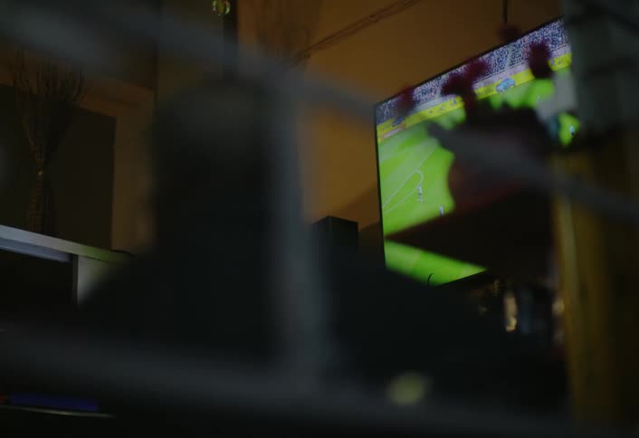 The video shows the back of a persons head partially obscured by blurry foreground elements as they sit in front of a brightly lit television displaying a soccer match the room appears dimly lit emphasizing the glow of the screen