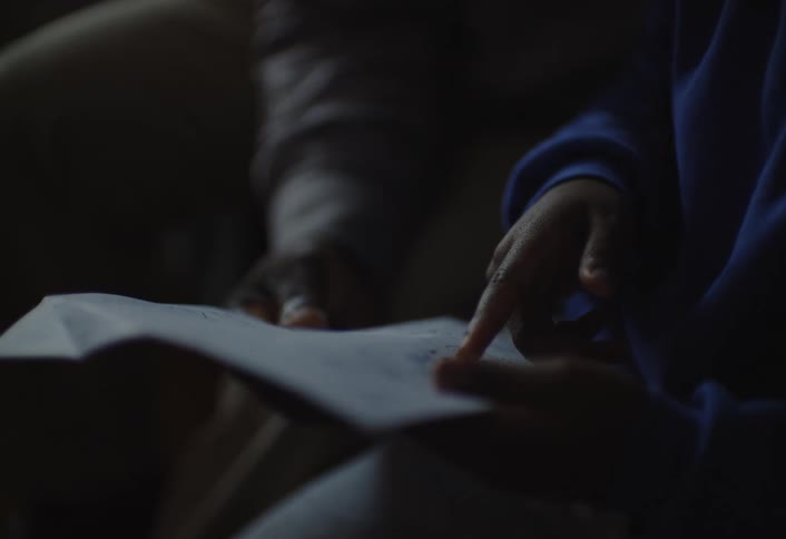 The video features a closeup shot of two peoples hands one set of hands wearing a longsleeved gray top is positioned behind a piece of white paper while the other set wearing a blue longsleeved shirt points to something on the paper