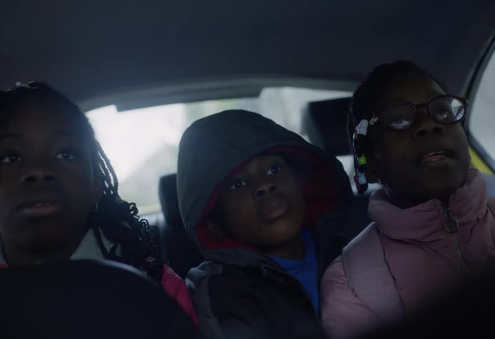The video shows three children in the back seat of a car looking forward the child in the middle is wearing a dark hooded jacket while the child on the right has glasses and is wearing a pink jacket