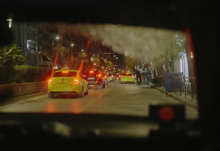 From inside a vehicle at night a long line of yellow taxis and other cars are visible on a city street with lights reflecting on the trees along the sidewalk