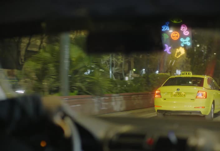 A yellow taxi drives through a lively city street at night flanked by glowing storefronts neon signs and illuminated highrise buildings