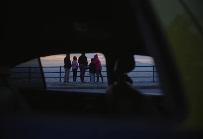 People by a waterfront railing at sunset seen through a car window