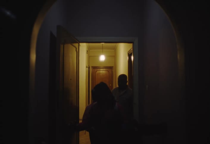 A man walks in opening the door as the light comes on then walks out of a room into a hallway at night
