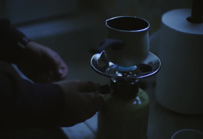 A person prepares coffee using a small stovetop setup in dim lighting