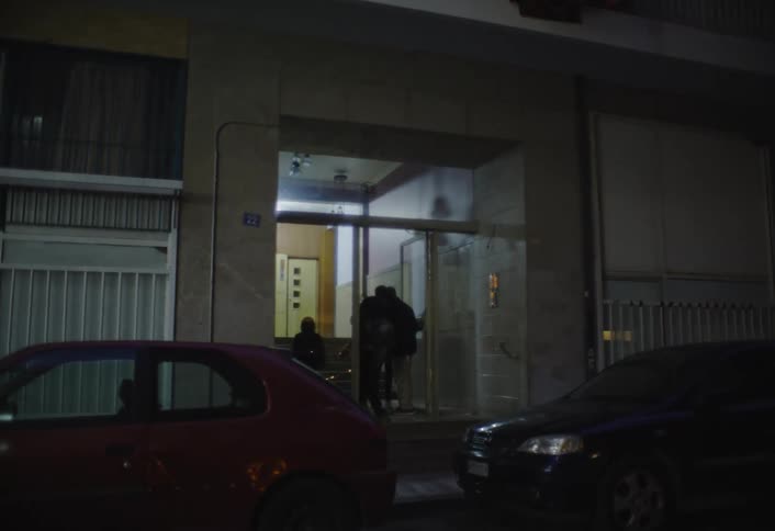 The video shows a building entrance at night with two people in red and pink jackets walking towards it a red car is parked on the left side and a darker car on the right