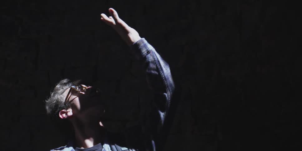 The video features a person in a dimly lit setting their face illuminated by an unseen light source from below they are looking upwards with one arm raised