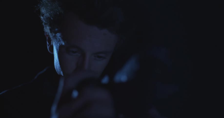 A man intently examines something in dim blue lighting his face partially illuminated in the darkness