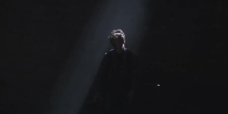 A young man staring up at a stage light that briefly reveals him