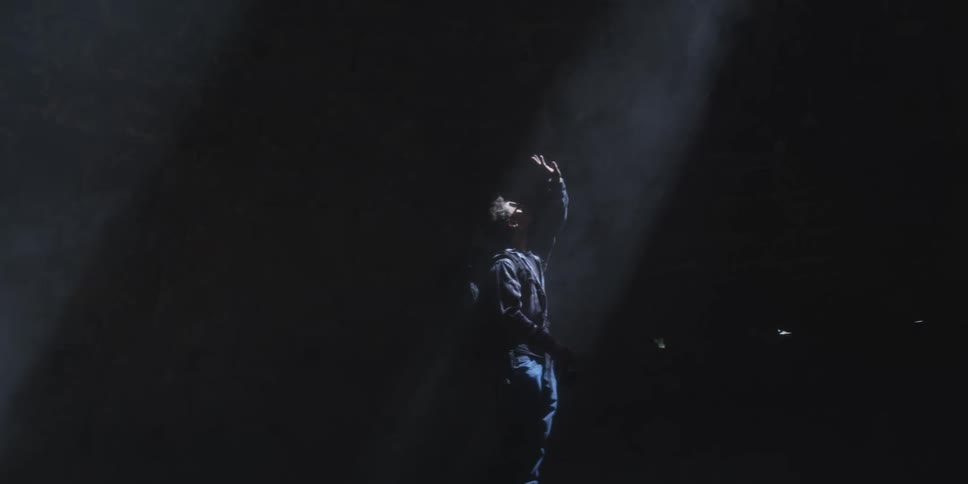 A man stands in a dark room reaching upward with one hand while aiming a camera at the ceiling under a slanted beam of light