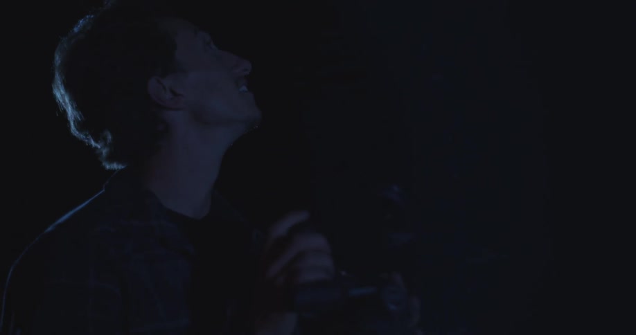 A young man is outdoors at night looking through his camera when he suddenly begins glancing left and right at the sky in fear