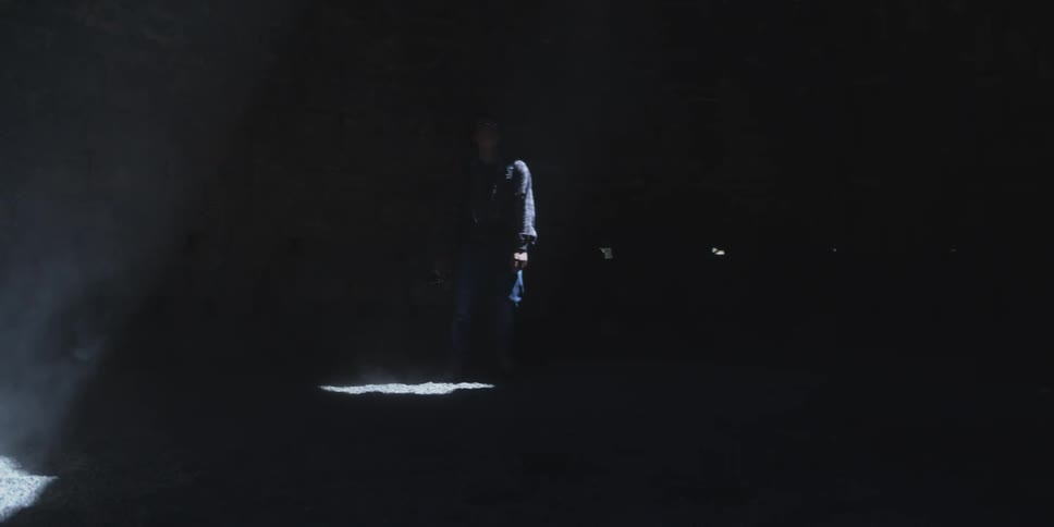 A person stands in a dark room under a single beam of light holding a camera up to their face