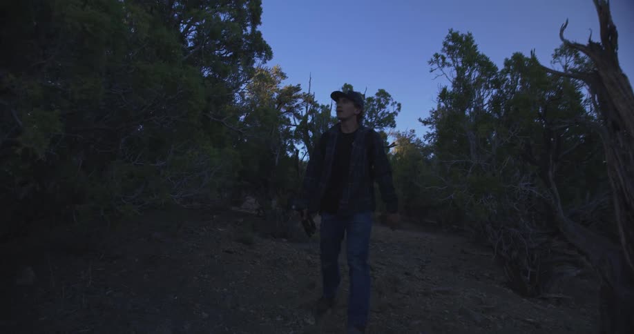 A young man wearing a hat walks along a trail in the woods at sundown