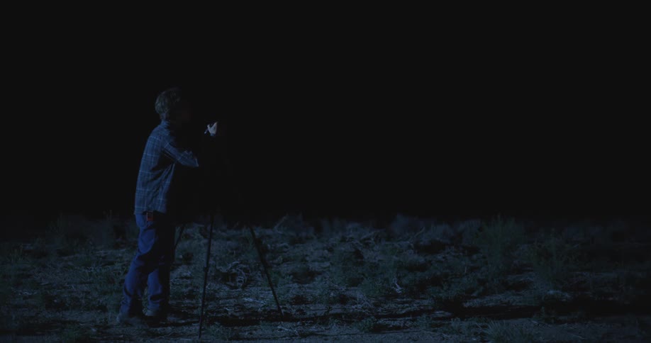 A person stands in a dark outdoor area operating a tripod mounted camera with surrounding trees faintly visible