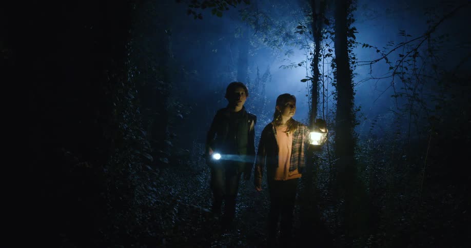 Two teens are walking in the woods at night the girl is holding a lamp and the boy is holding a torch as they walk slowly looking scared