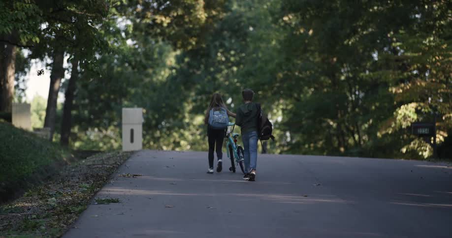 Girl walking with a bicycle and a boy following behind with a backpack together on a city street