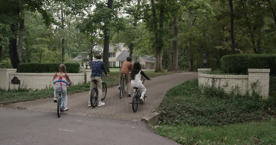 Four children pedal along a tree lined path toward a large house nestled in dense foliage