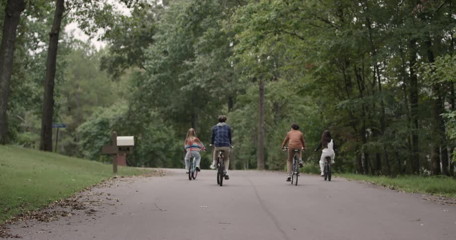 Four teenagers are riding bikes on a lonely road in a suburban area surrounded by trees at midday