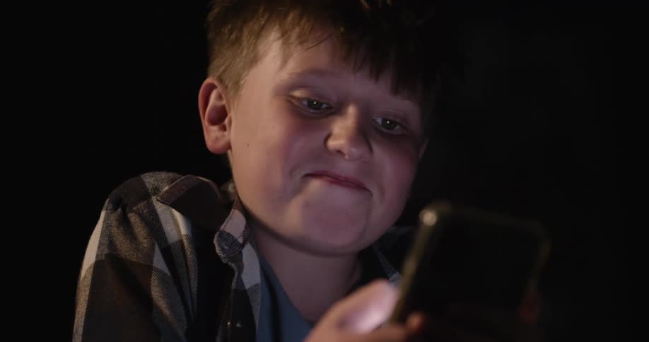 A boy in a plaid shirt uses a smartphone in a dimly lit space