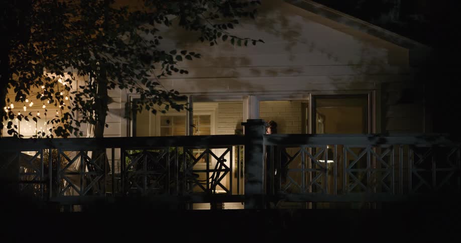 A teenage boy walks out of the front porch of a house and rests on the rail fence in front of the building