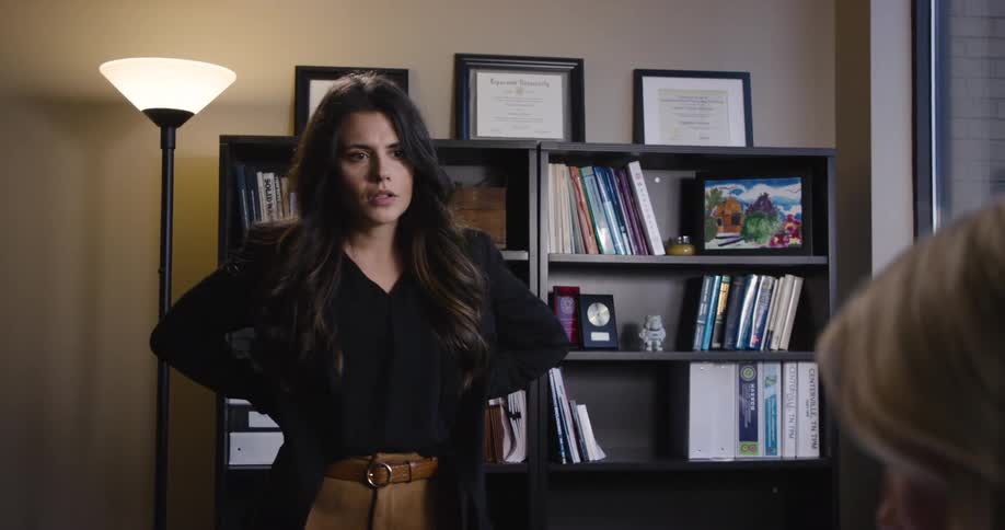 A woman stands in an office with a serious expression across from a person framed by bookshelves and diplomas behind her