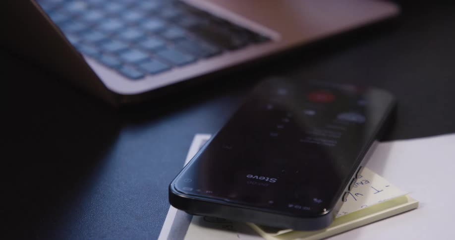 A smartphone rests on a stack of sticky notes beside a laptop on a dark desk as a person types on a laptop