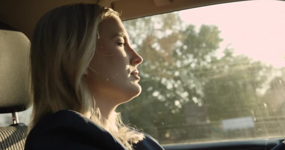 A blonde woman drives through a suburban neighborhood appearing content as morning light filters in
