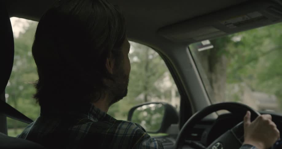 A man in a plaid shirt smiles while driving a car through a green suburban area