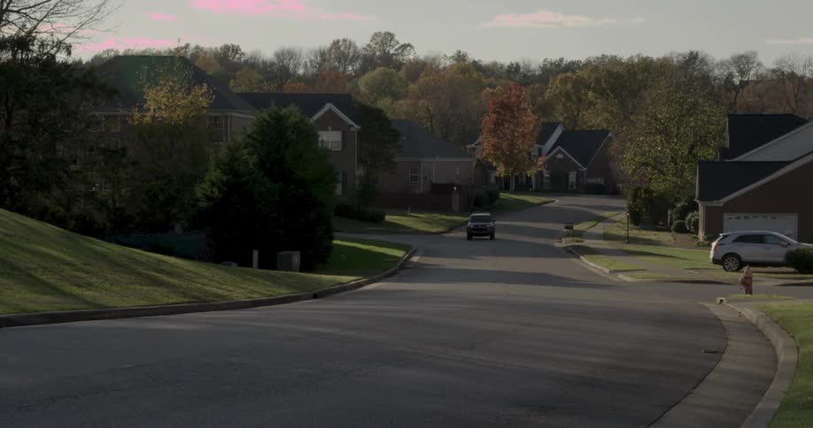 A quiet suburban neighborhood street gently slopes downhill in soft evening light