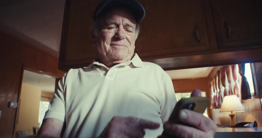 The video features an elderly man wearing a lightcolored collared shirt and a baseball cap holding a smartphone he appears to be engaged in a video call or recording a message as he smiles and interacts with the camera