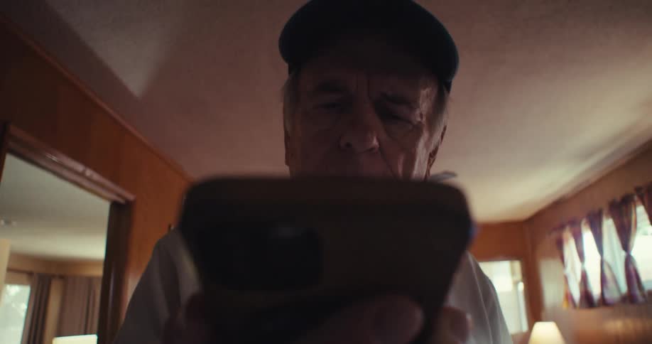 The video features a closeup of an elderly man with a blue veteran cap looking directly at the camera while holding a smartphone the background is a warmtoned interior with light wooden walls and a fan visible on the ceiling