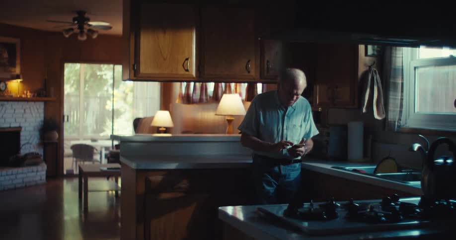 An elderly man stands in a dimly lit kitchen holding an object with the living room softly lit behind him