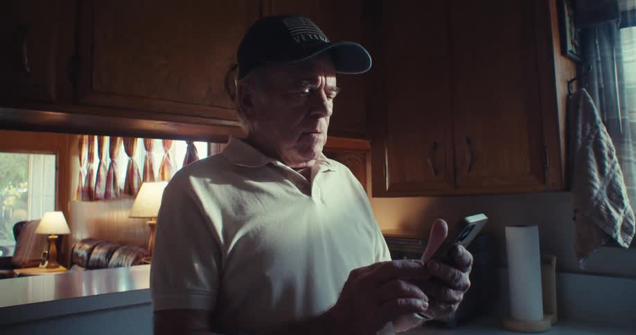 Elderly man in cap standing in kitchen interacting with smartphone