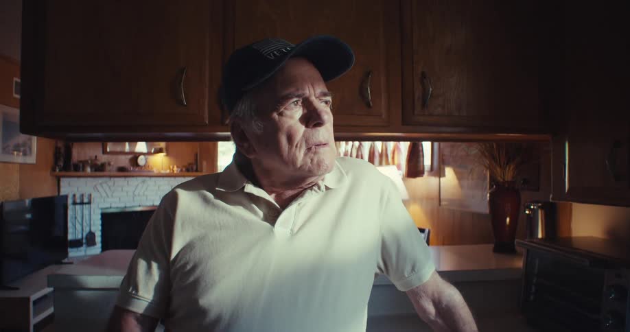 The video features a man wearing a veteran cap and a white polo shirt standing in a kitchen with wooden cabinets he is holding a smartphone and looking at the screen with sunlight streaming in from a window in the background