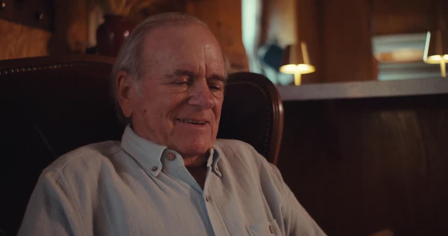 The video features a closeup shot of an elderly man with a lightcolored collared shirt sitting in a leather armchair he is looking at and interacting with a smartphone and seems to be smiling