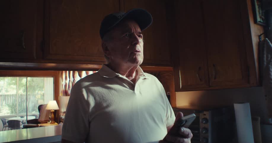 A man stands in a kitchen using his phone appearing slightly confused or thoughtful