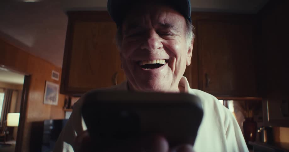 The video features a blurred closeup of an elderly man holding a smartphone with a veteran baseball cap visible he appears to be talking and laughing while looking at the screen