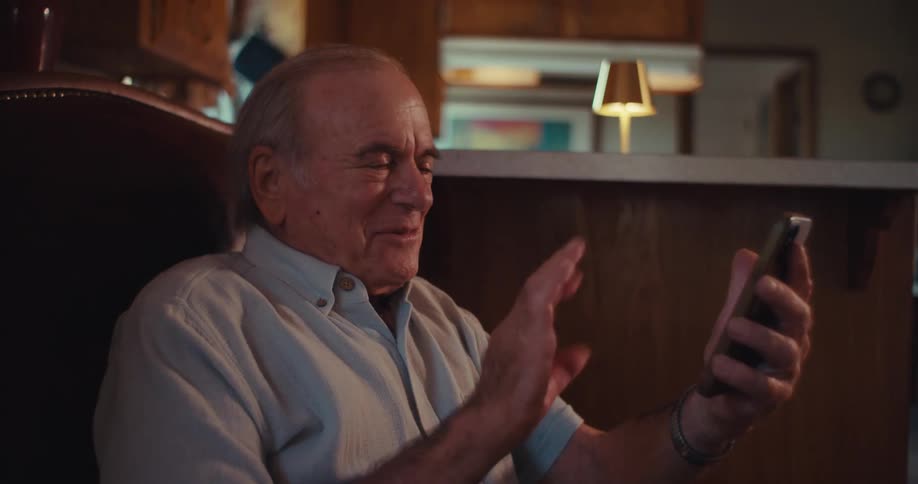 The video features an elderly man sitting in a comfortable armchair interacting with his smartphone he smiles and waves at the camera as if engaged in a video call or recording a message