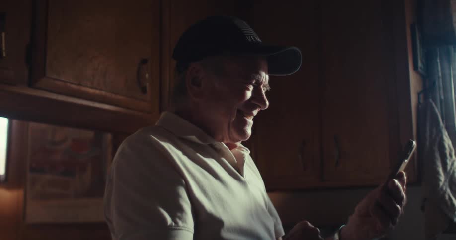The video shows a man wearing a baseball cap and a lightcolored shirt standing in a kitchen or dining area he is looking at his phone and appears to be interacting with it