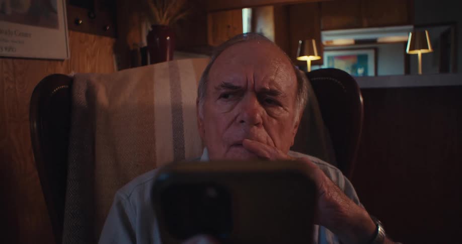 An elderly man sitting on a chair in his living room looking at his smartphone