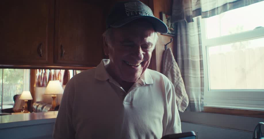 The video shows a man wearing a veteran cap and a white polo shirt interacting with a smartphone he smiles and gestures with his hands indicating he is engaged in a video call or viewing content on the device