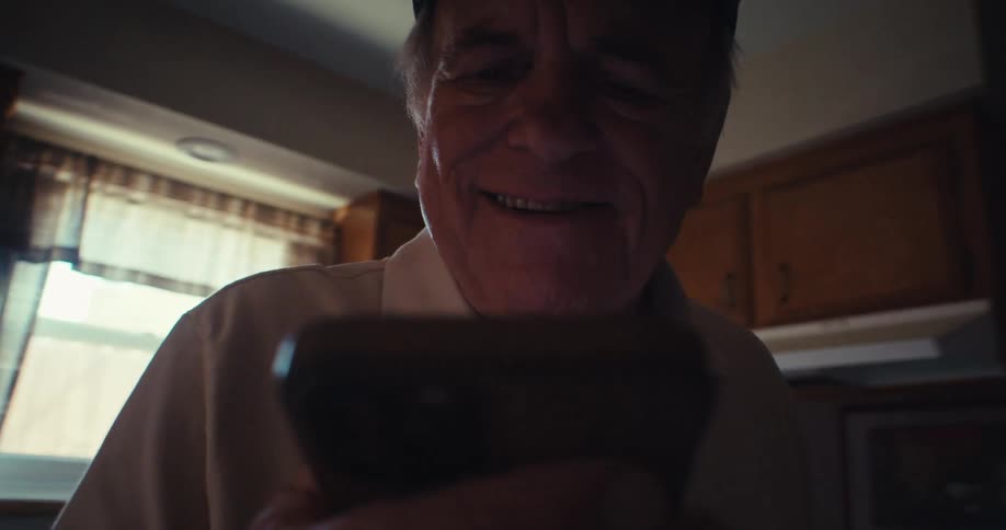 An older man smiles warmly while looking at his phone in a dimly lit kitchen
