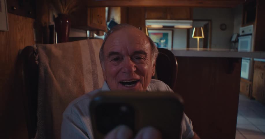 An elderly man smiles during a video call in a cozy kitchen