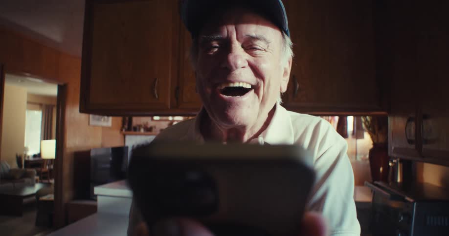 An elderly man wearing a cap is laughing in his kitchen while talking on the smartphone