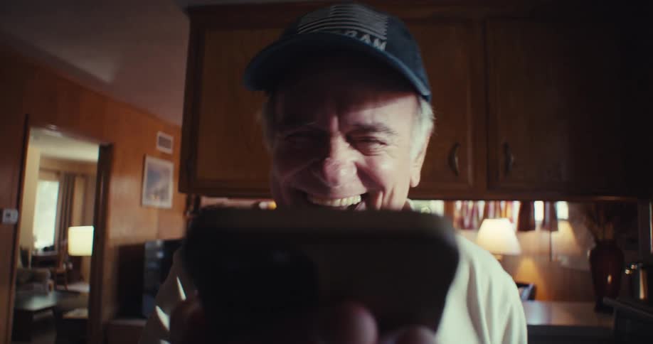 The video features an elderly man wearing a veteran baseball cap looking directly at the camera with a wide joyful smile he appears to be laughing heartily possibly in reaction to something on a smartphone which is held out of frame