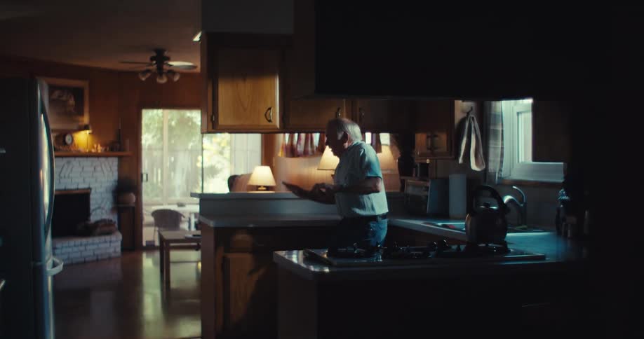 The video shows an elderly man in a kitchen setting holding a smartphone he is looking at the phone and appears to be laughing or reacting with amusement to something he sees or hears