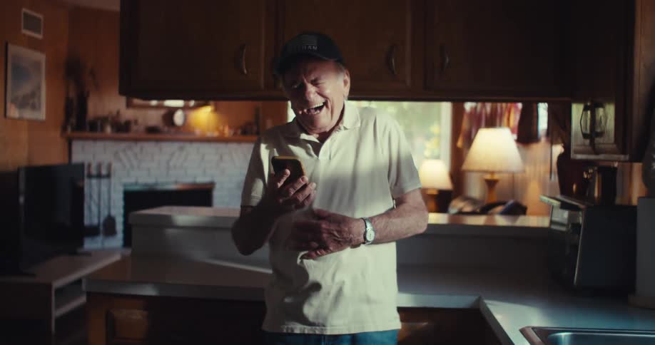 Elderly man laughing while standing in the kitchen and holding a smartphone